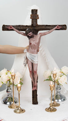 May include: A detailed sculpture of Jesus on a wooden cross. The figure is depicted with a crown of thorns and a white loincloth. The cross is brown, and the figure has a realistic skin tone. The scene includes lit candles and white roses.