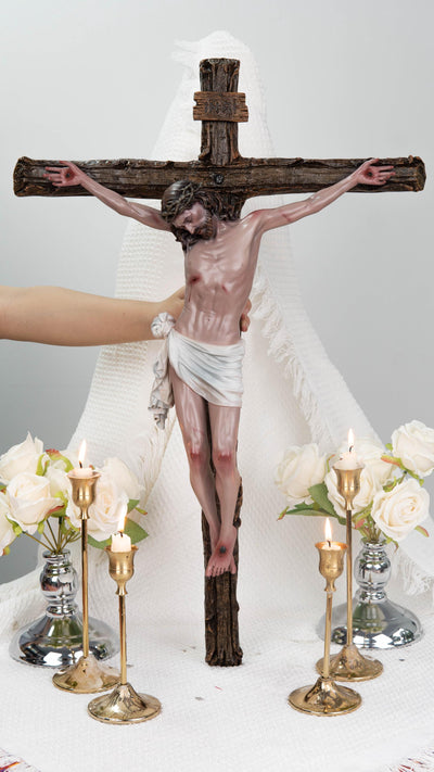 May include: A detailed sculpture of Jesus on a wooden cross. The figure is depicted with a crown of thorns and a white loincloth. The cross is brown, and the figure has a realistic skin tone. The scene includes lit candles and white roses.